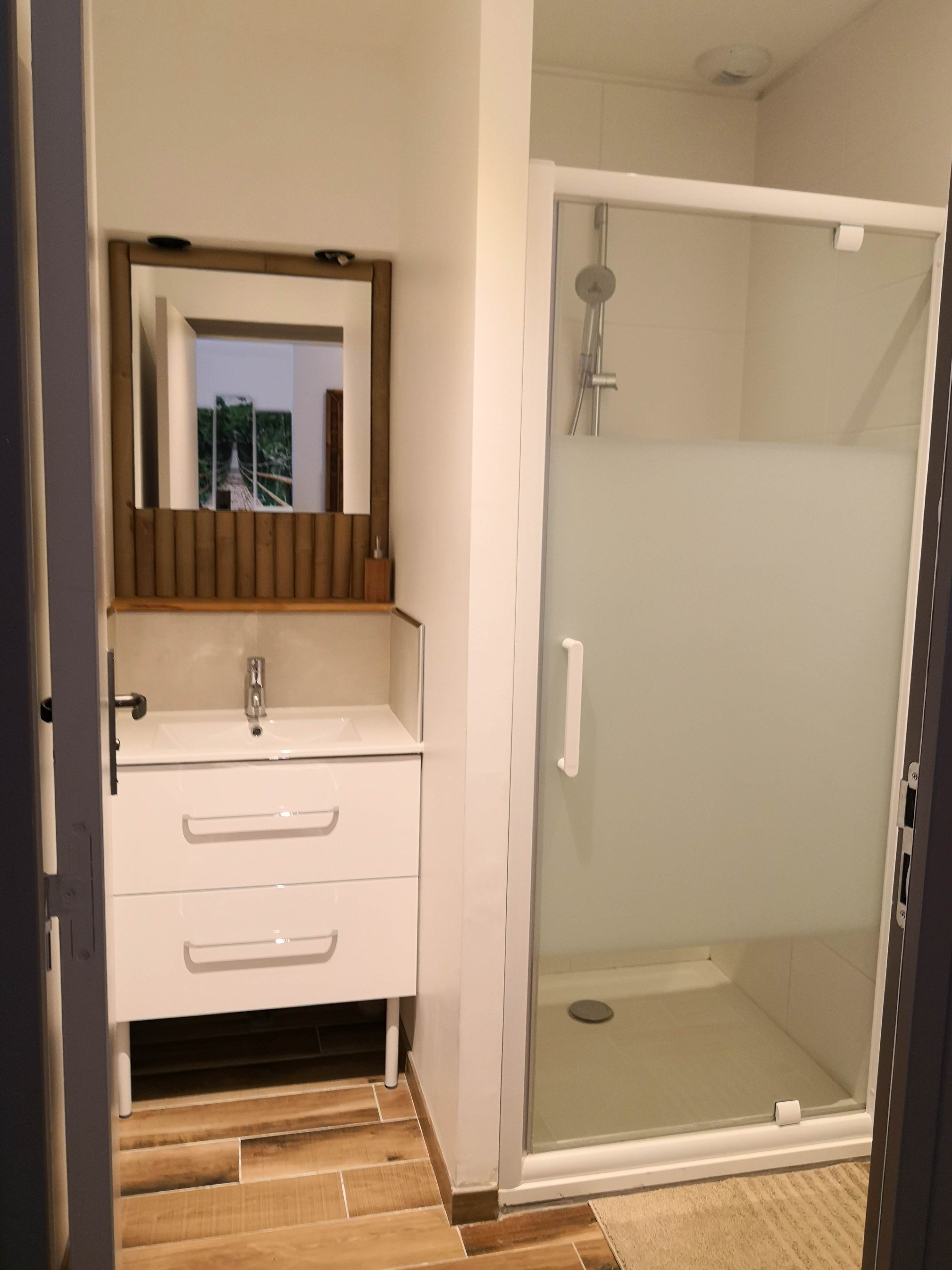 Salle de bain avec vasque blanche sur meuble à tiroirs, miroir au cadre de bambou et douche à porte en verre dépoli.
