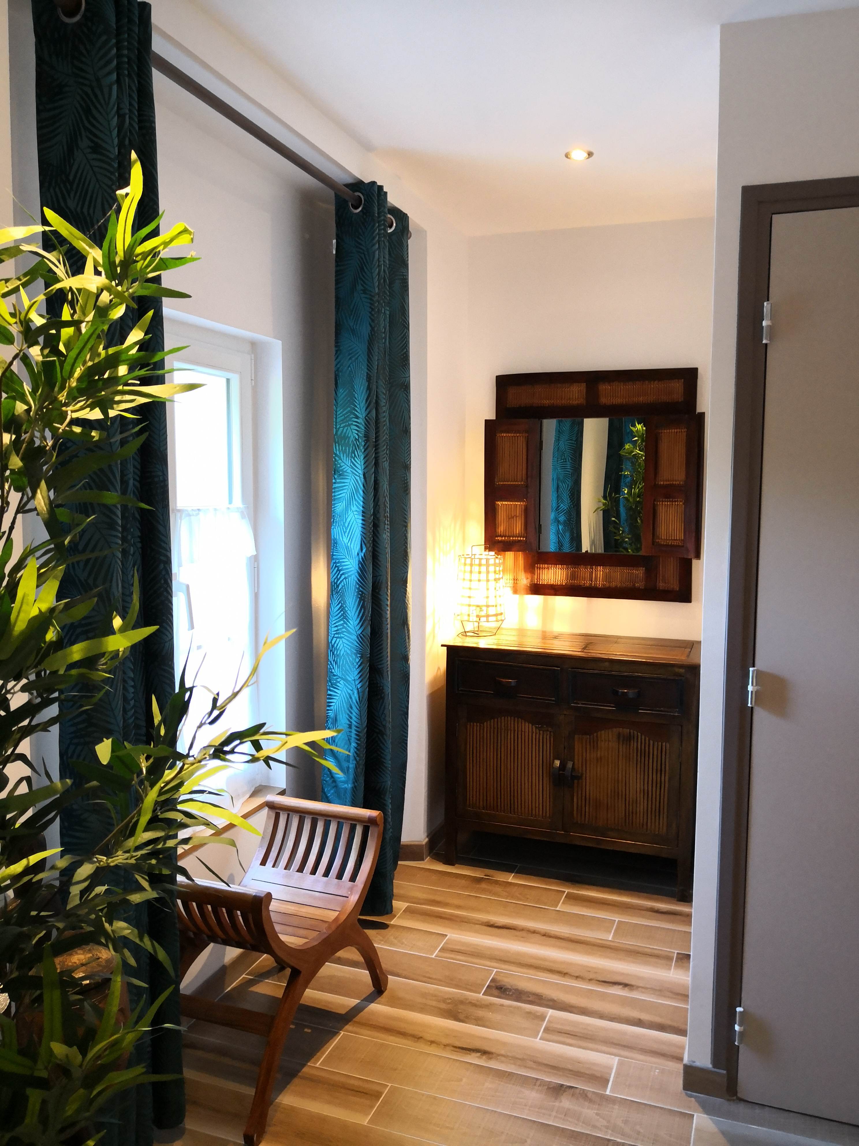 Entrée de la chambre avec rideaux à motif de palmes turquoise, banc et commode en bois exotique sous un miroir à volets de bambou.