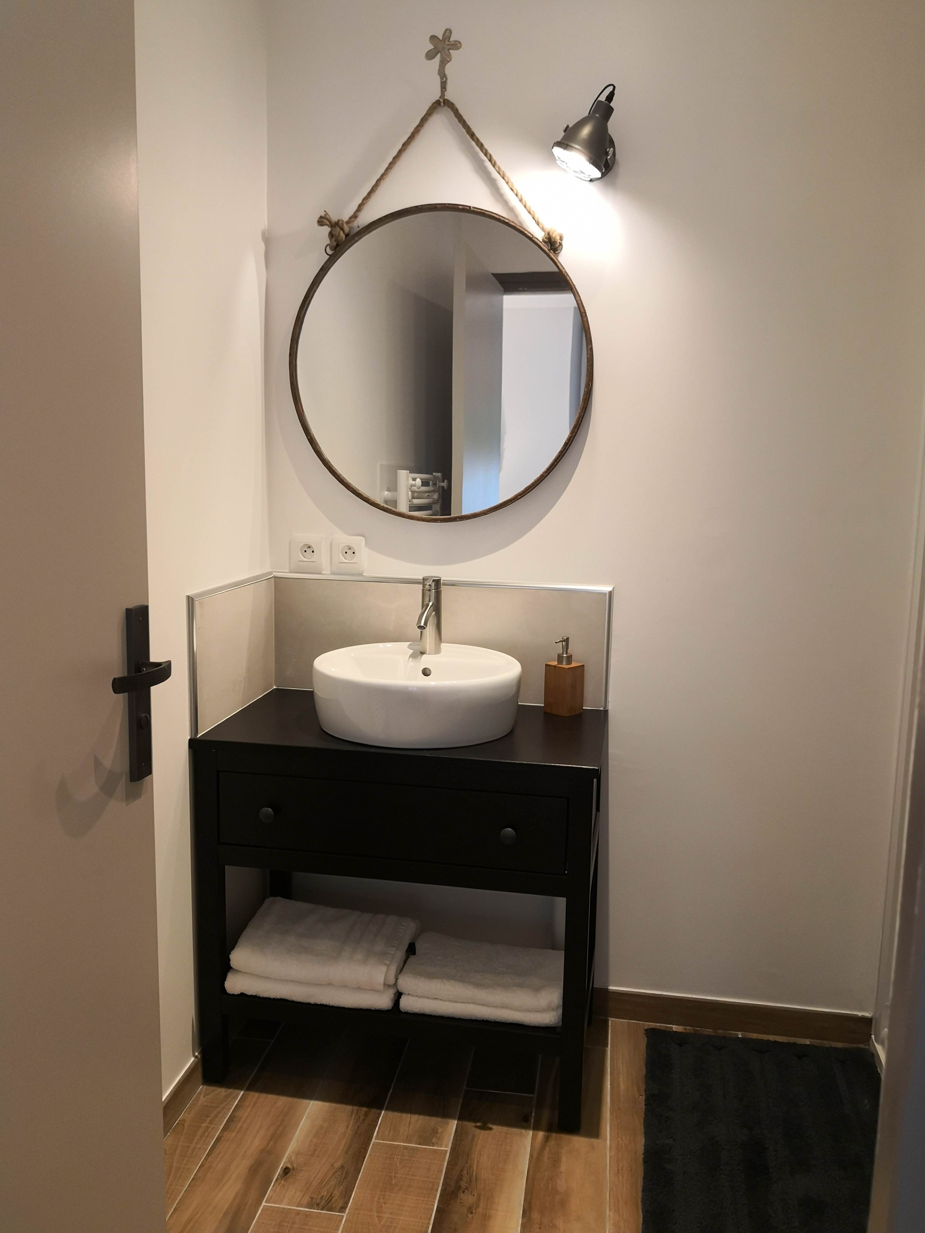 Salle de bain avec vasque blanche sur meuble noir, miroir rond suspendu par une corde et serviettes blanches pliées.